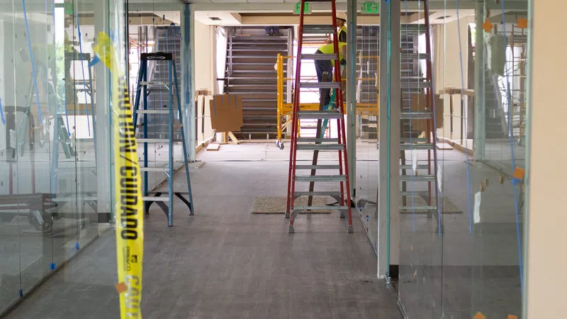 Interior office space under construction with glass walls, ladders, caution tape, and worker on a ladder.