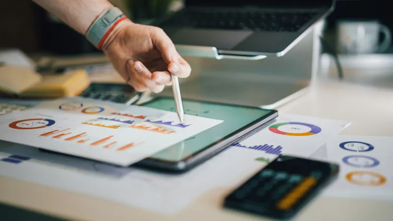 Person using a stylus on a tablet displaying financial charts and graphs on a busy desk.