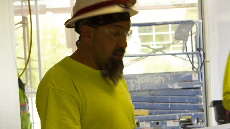Construction worker in a bright yellow shirt and hard hat reviewing documents on a clipboard indoors.