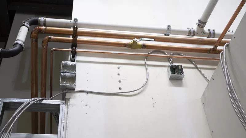 Copper and PVC pipes installed along a white wall with metal conduit wiring and junction boxes in a building under construction.