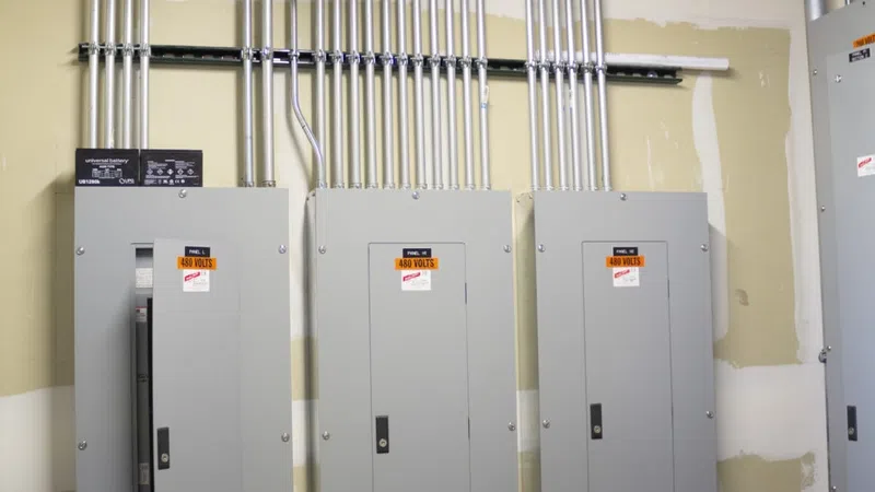 Three gray industrial electrical panels with 480 volts warning labels mounted on a beige wall with metal conduit pipes above.
