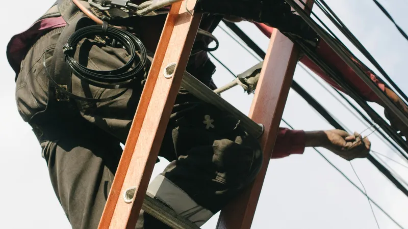 Technician climbing a ladder to repair or install cables on an electrical pole with numerous wires.