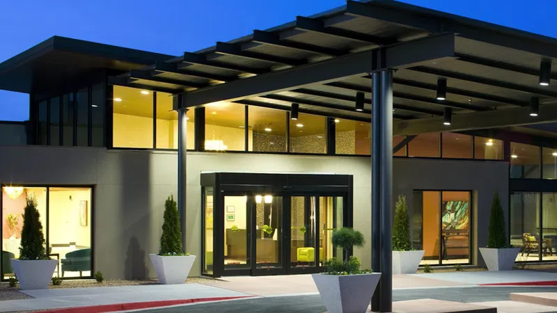 Modern building entrance with large glass doors, potted plants, and covered walkway illuminated at dusk