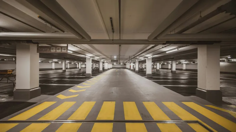 Empty underground parking garage with yellow pedestrian stripes and white support pillars under ceiling pipes.