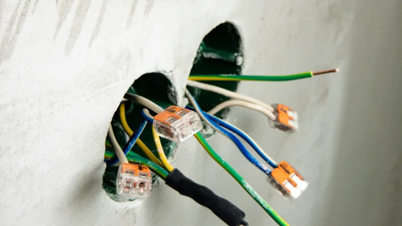 Exposed electrical wiring with orange connectors protruding from holes in a white plastered wall.