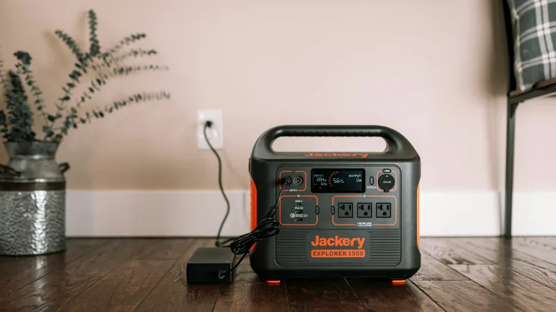 Jackery Explorer 1500 portable power station on wood floor plugged into wall outlet with plant and chair nearby