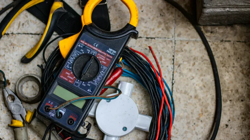 Digital clamp meter, coiled electrical wires, and pliers on tiled floor for electrical work.