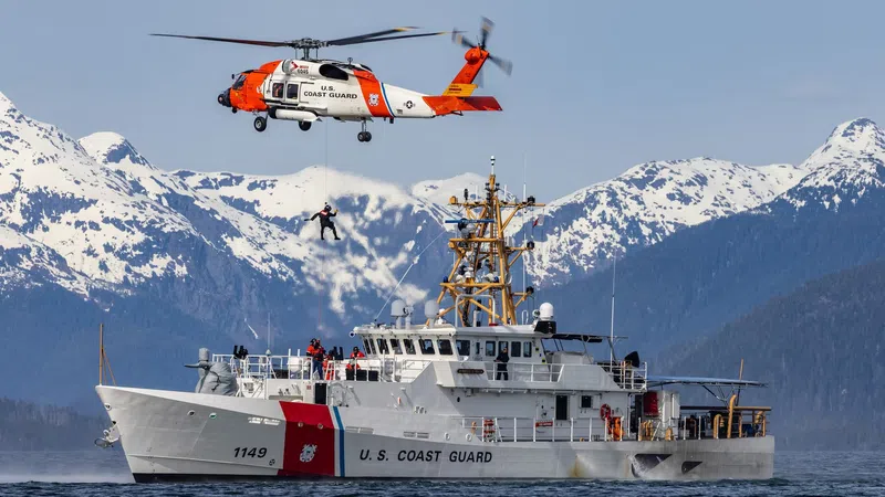 U.S. Coast Guard helicopter performing a rescue hover over a Coast Guard cutter with snowy mountains in the background