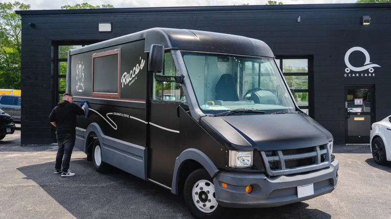 Black food truck named Rocco's parked outside a black building with a person cleaning its side on a sunny day.
