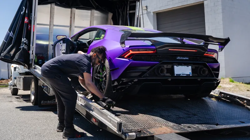 Purple Lamborghini Huracan being secured on a flatbed tow truck by a worker in gloves.