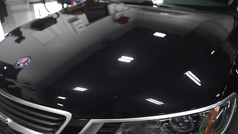 Close-up of a shiny black car hood with a Saab emblem, grille, and headlight under bright indoor lights