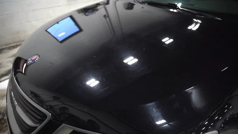 Close-up of a black car hood with reflections of overhead lights and a Saab logo.
