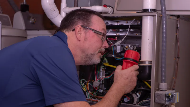 Technician with glasses inspecting and repairing HVAC system wiring using a red flashlight indoors.