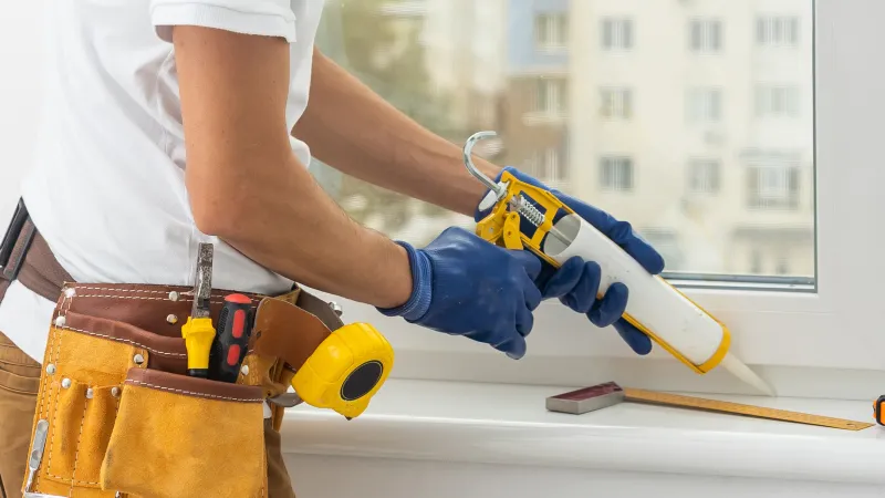 Construction worker applying sealant to window frame with caulking gun while wearing blue gloves and tool belt.