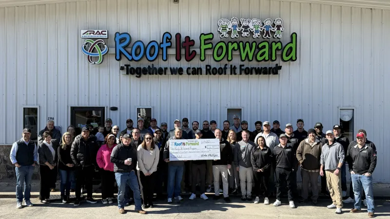 Group of Roof It Forward team members standing outside building holding a large check for a charity event.