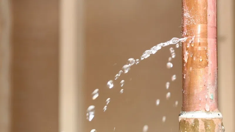 Close-up of a copper pipe leaking water in a steady stream through a small hole in the pipe.