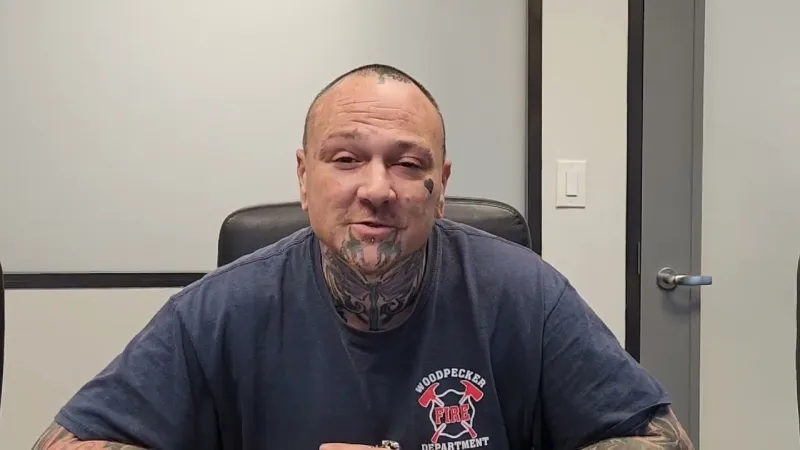 Man with facial and neck tattoos sitting in a chair wearing a Woodpecker Fire Department shirt indoors.