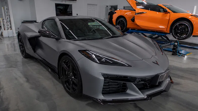 Matte gray sports car in showroom with bright orange car on lift in background.