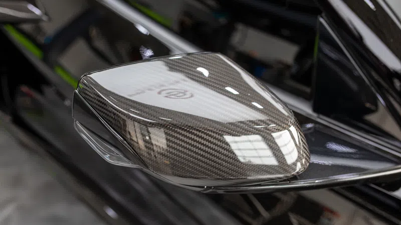 Close-up of a carbon fiber car side mirror with black glossy finish and clear turn signal light.