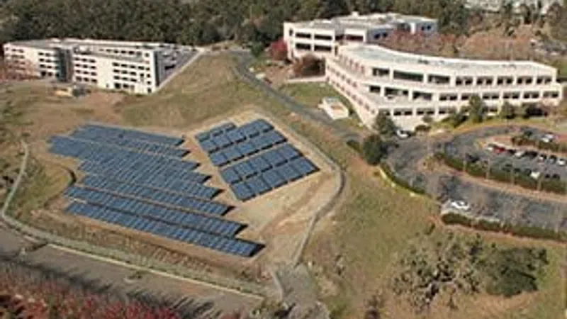 Aerial view of solar panels installed on a sloped area near office buildings surrounded by trees and parking lots.