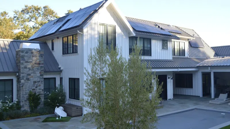 Modern white house with solar panels on roof, stone chimney, large windows, and spacious backyard patio.