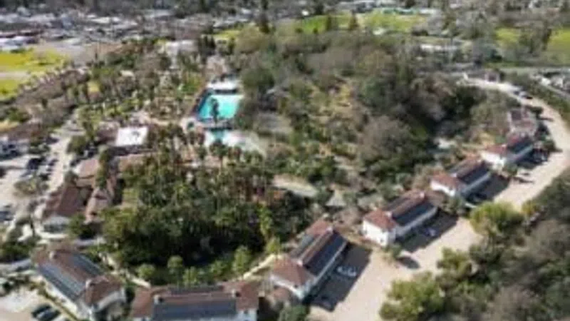 Aerial view of a resort with multiple buildings, a pool, parking areas, and surrounding greenery and hills.