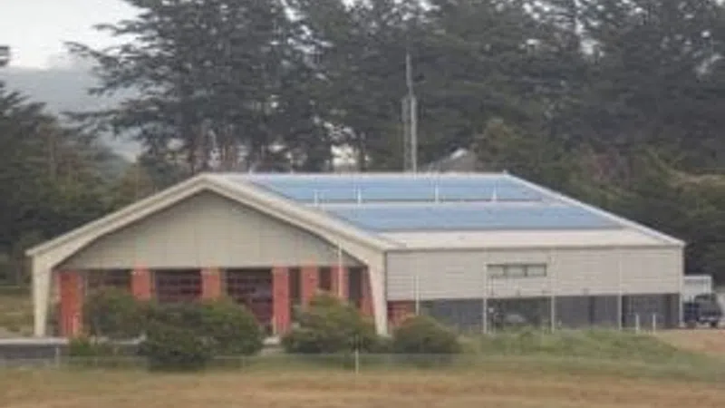 Large industrial building with solar panels on the roof surrounded by trees and grassy field.