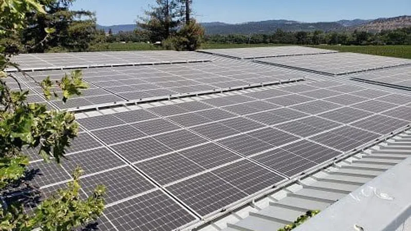 Solar panels installed on a rooftop with trees and hills visible in the background under clear skies.