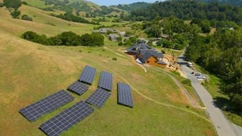 Solar panels installed on a grassy hillside near a rural building complex under clear sky.