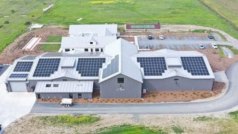 A large farm building with solar panels on the roof surrounded by green fields and a gravel road.