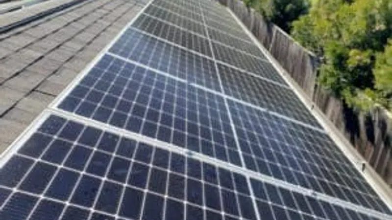 Solar panels installed on a rooftop in bright sunlight with trees and fence in the background.