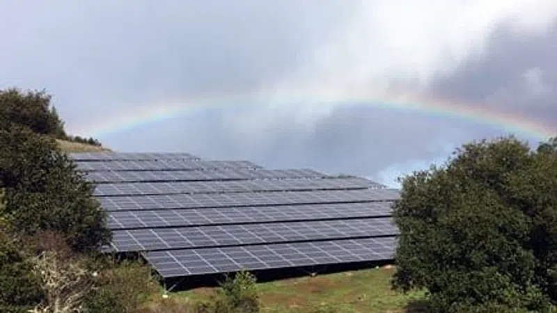 Solar panels on a green hillside with a rainbow and cloudy sky in the background.