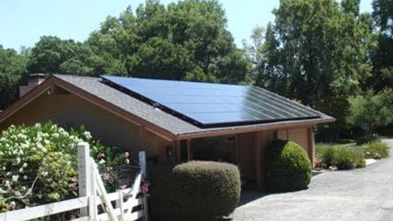 Residential house with solar panels on the roof surrounded by greenery and a white fence under clear blue sky.