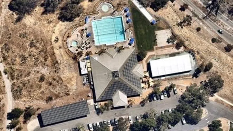 Aerial view of a community center with a large swimming pool, parking lot, and surrounding dry landscape.