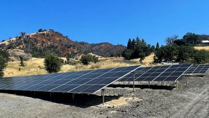 Large solar panel array installed on a dry hillside under a clear blue sky with mountainous background.