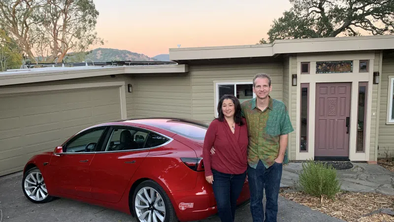 Couple standing beside a red Tesla Model 3 parked in front of a modern house at sunset.