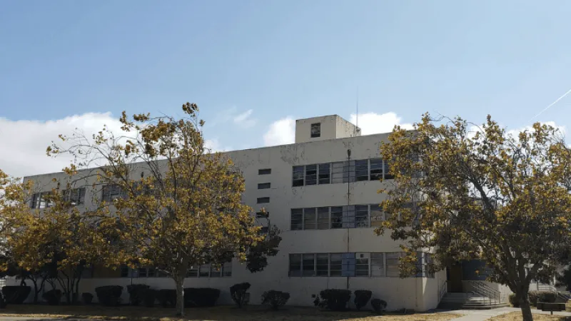 Three-story beige building with multiple windows surrounded by trees with yellow leaves under a clear blue sky