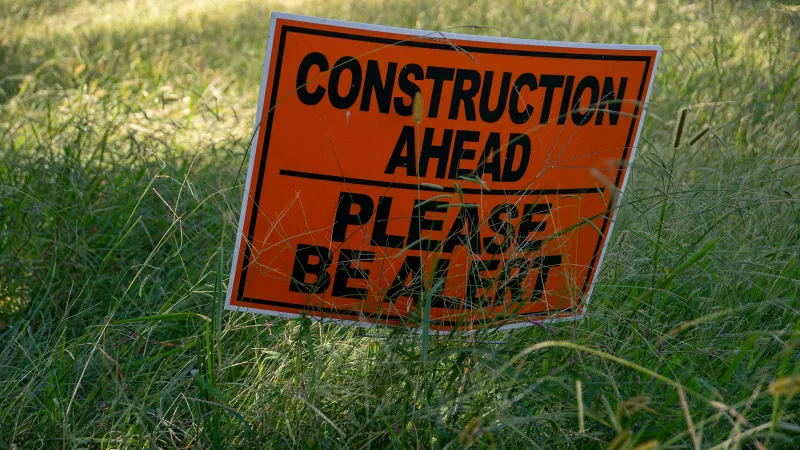 Orange construction warning sign in tall grass reading Construction Ahead Please Be Alert.