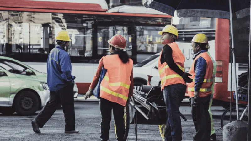 Four construction workers wearing safety helmets and reflective vests standing near vehicles on a city street.