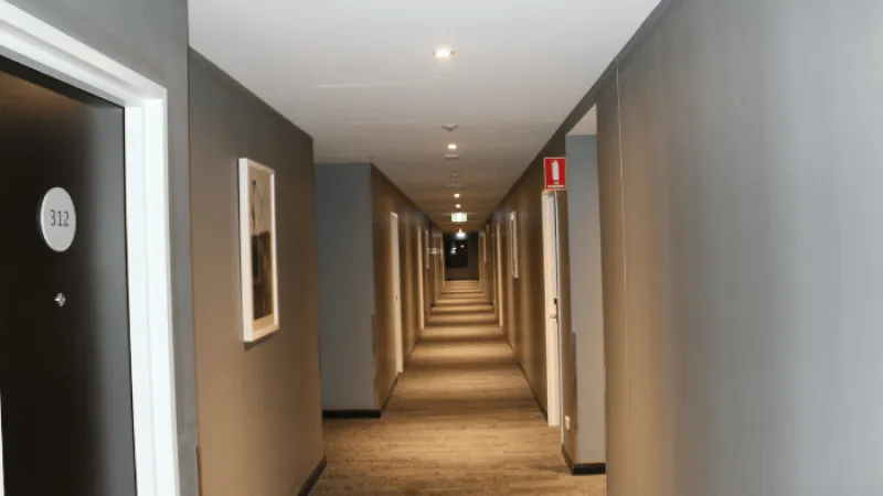 Long hotel hallway with carpeted floor, multiple doors numbered, framed artwork, and ceiling lights.