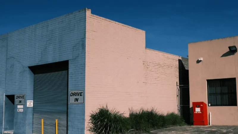 Industrial building with drive-in garage doors, yellow bollards, green shrubs, and a red fire hose box under clear blue sky.