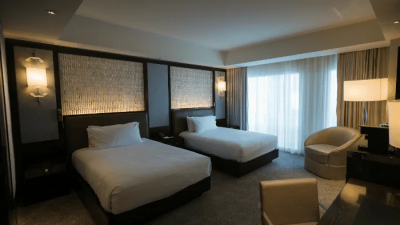 Modern hotel room with two beds, upholstered chair, desk, soft lighting, and large window with curtains.