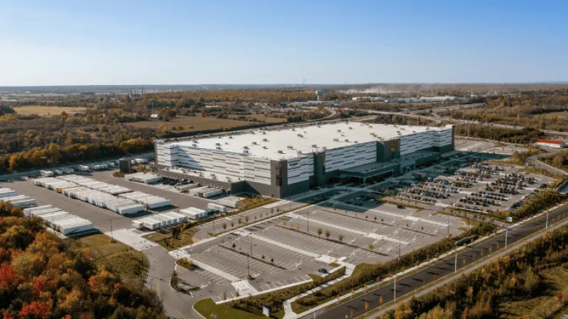 A large industrial warehouse with parking lots and semi-trailers surrounded by trees under a clear sky.