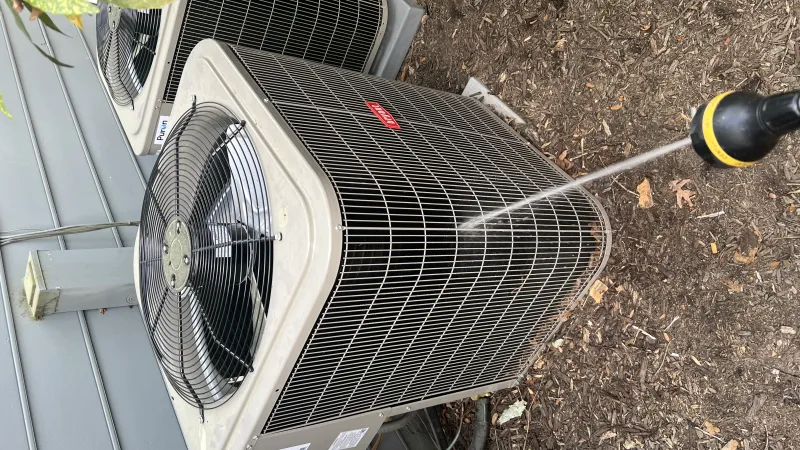 Pressure washing an outdoor air conditioning unit with a hose on garden soil beside a house siding.
