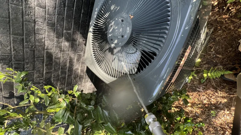 Person cleaning outdoor HVAC unit with a water hose surrounded by green plants and brick wall.