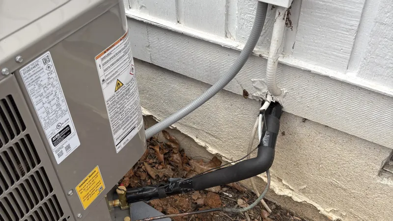 Outdoor air conditioning unit connected to a white wooden house with insulated pipes and electrical wiring.