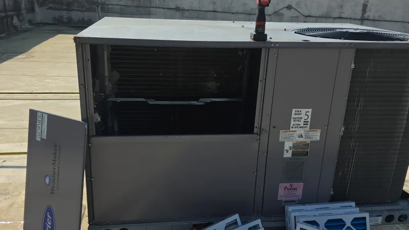 Open rooftop HVAC unit with removed air filters and a red cordless drill on top under clear sky.