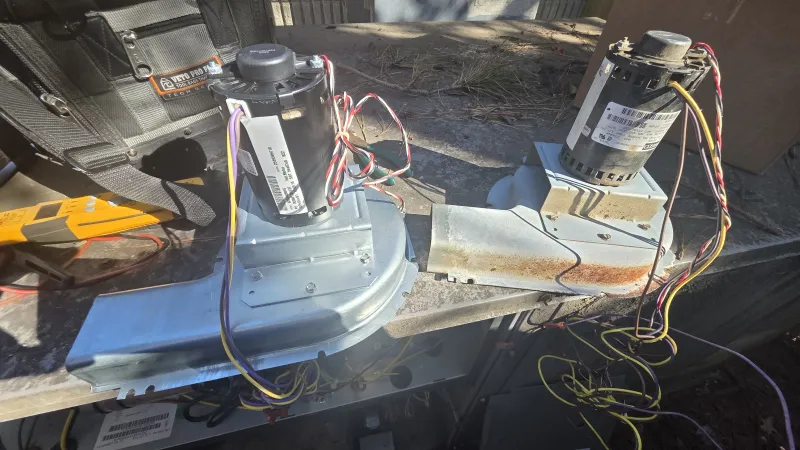 Two HVAC blower motors with wiring resting on a metal workbench in an outdoor setting.