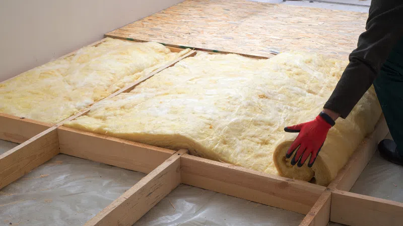 Worker installing yellow fiberglass insulation rolls on wooden floor frame for home thermal insulation.