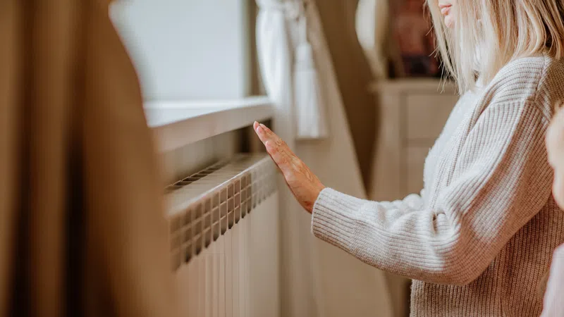 Woman in cozy sweater warming her hand on a home radiator near a window with curtains.